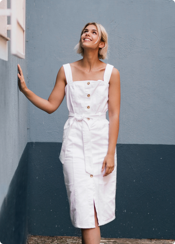 Mulher loira de vestido branco sorri e olha para cima, encostando na parede.