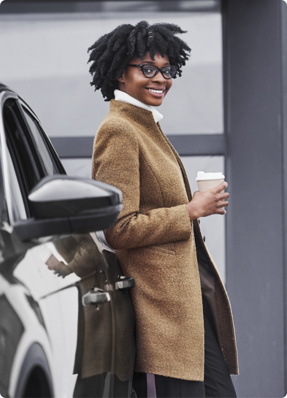 Mulher sorridente de óculos, com casaco marrom, encostada em um carro e segurando um café.