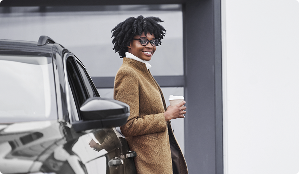 Mulher sorridente de óculos, com casaco marrom, encostada em um carro e segurando um café.