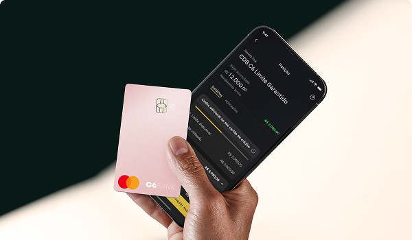 Mão segura um Cartão C6 Pink e um celular com o app C6 Bank.