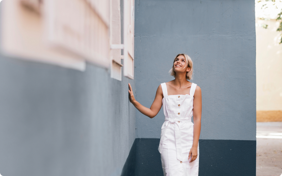 Mulher loira de vestido branco sorri e olha para cima, encostando na parede.