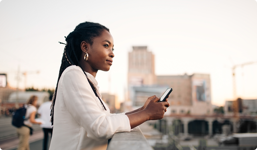 Mulher segura um celular e observa a cidade ao fundo.