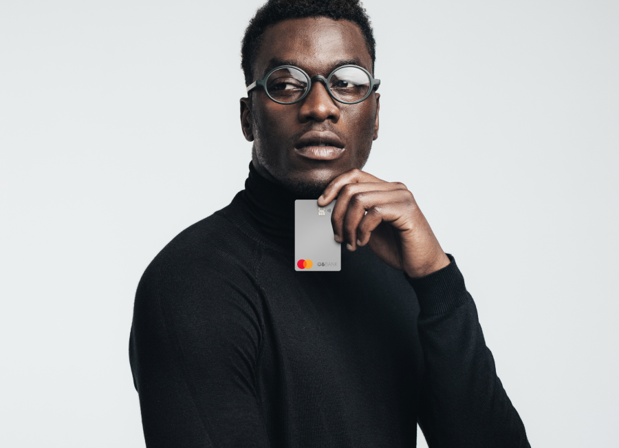 Foto de homem negro, de cabelos crespos, curto e preto. Ele está vestindo uma blusa preta de manga longa e gola alta e está segurando, com a mão esquerda e abaixo do rosto, um cartão do C6 Bank na cor prateada.
