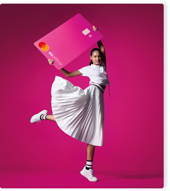 Menina levanta cartão rosa C6 Yellow ampliado em fundo vibrante. Visual jovem, alegre e cheio de movimento.