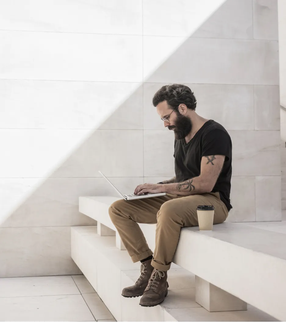 Homem barbado com tatuagens usa notebook sentado em um banco de concreto, com um café ao lado.