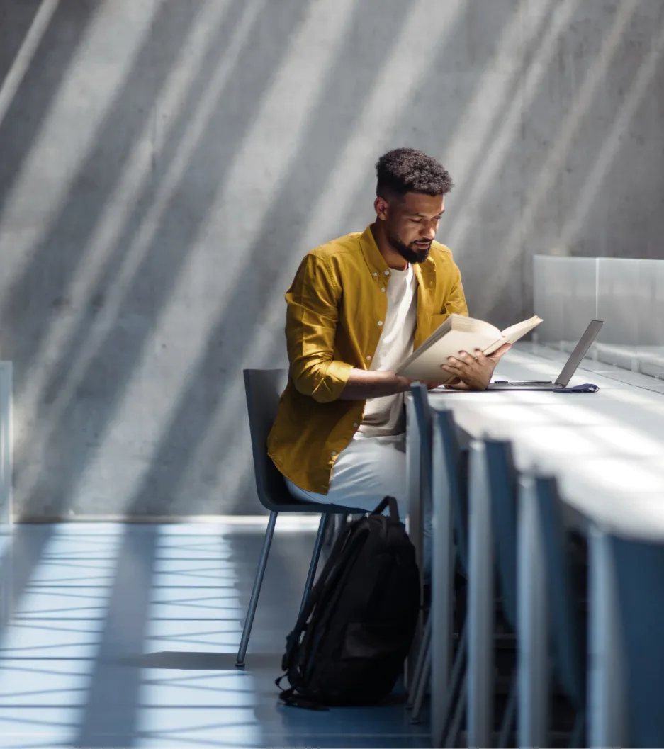 Homem com camisa mostarda lê um livro, com um notebook aberto e luz natural ao redor.