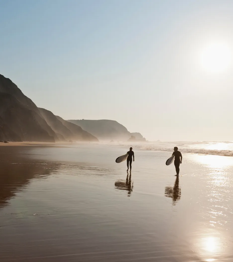 Dois surfistas caminham pela praia ao entardecer, com montanhas ao fundo e luz dourada do sol.