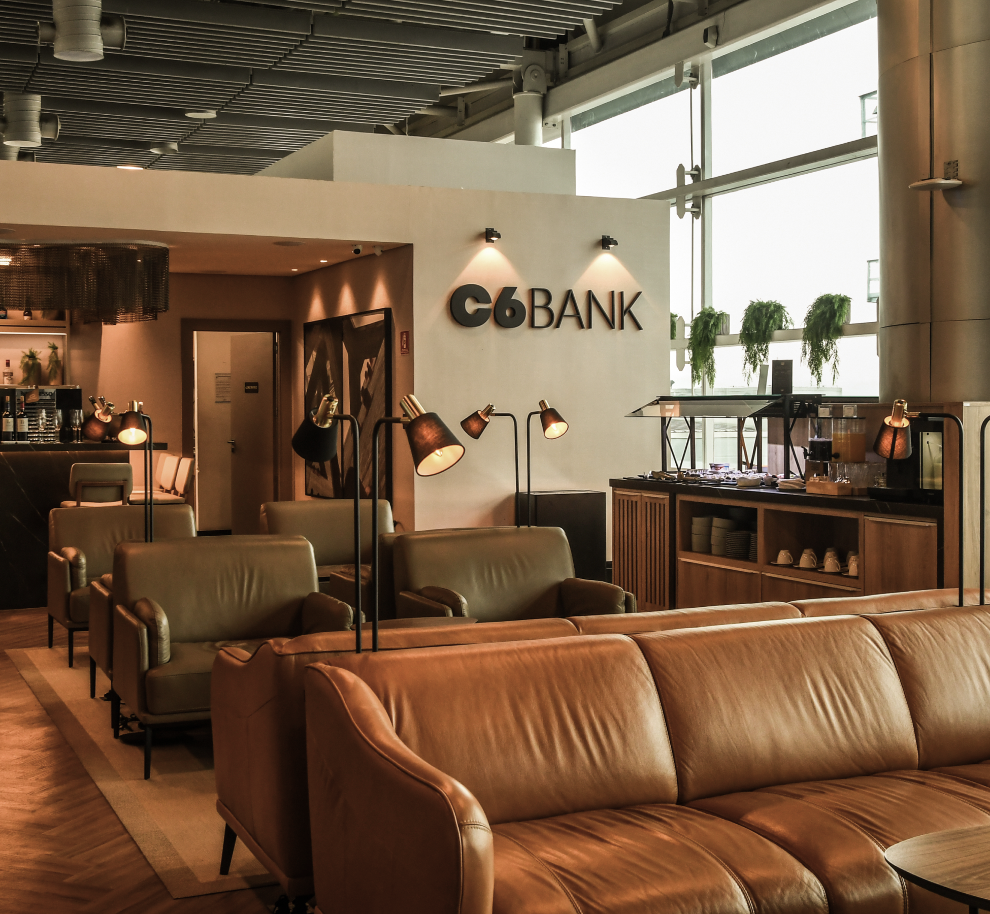 Sala VIP do C6 Bank com sofás de couro, buffet e logo da marca na parede em ambiente sofisticado.