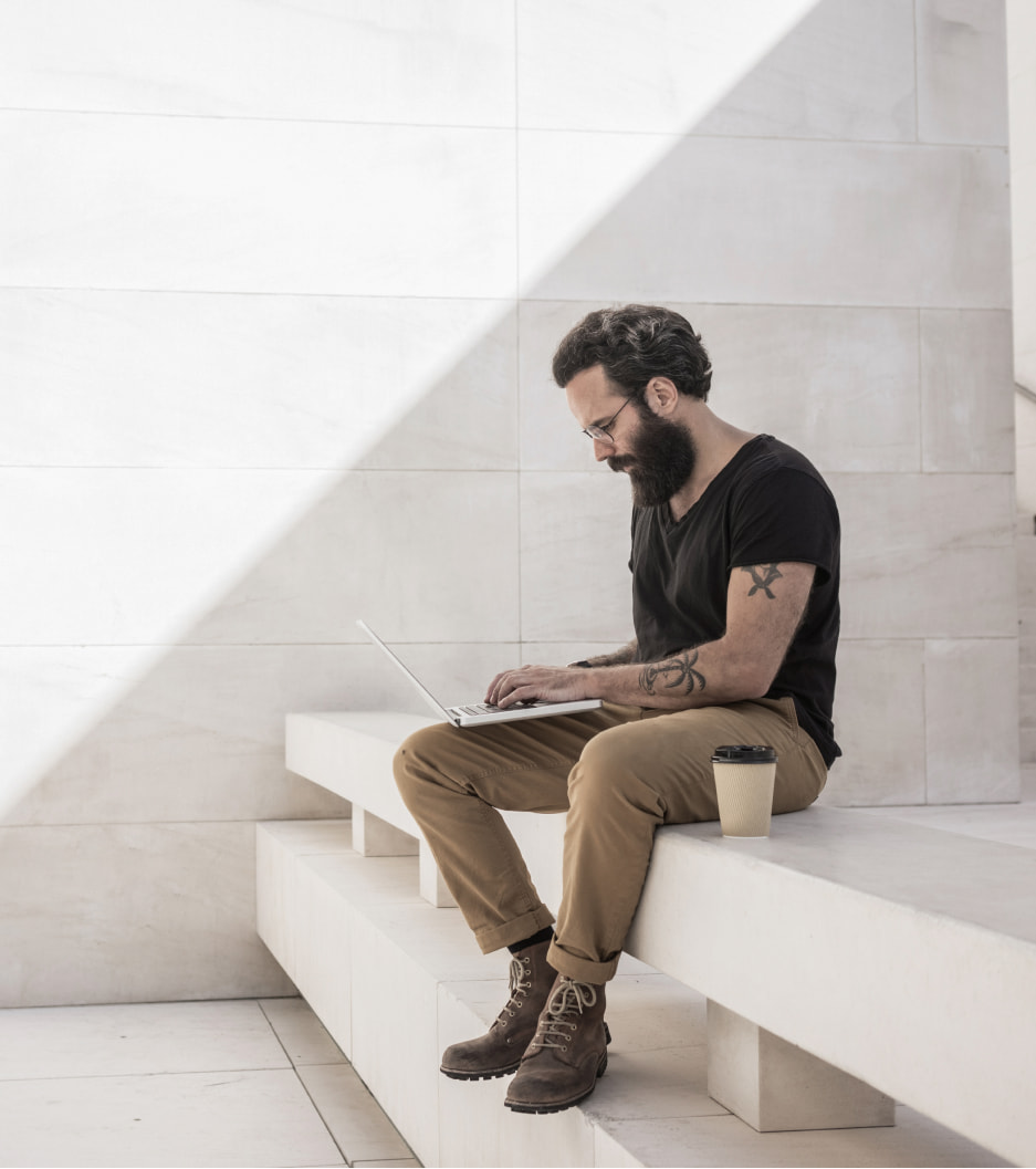 Homem barbado com tatuagens usa notebook sentado em um banco de concreto, com um café ao lado.