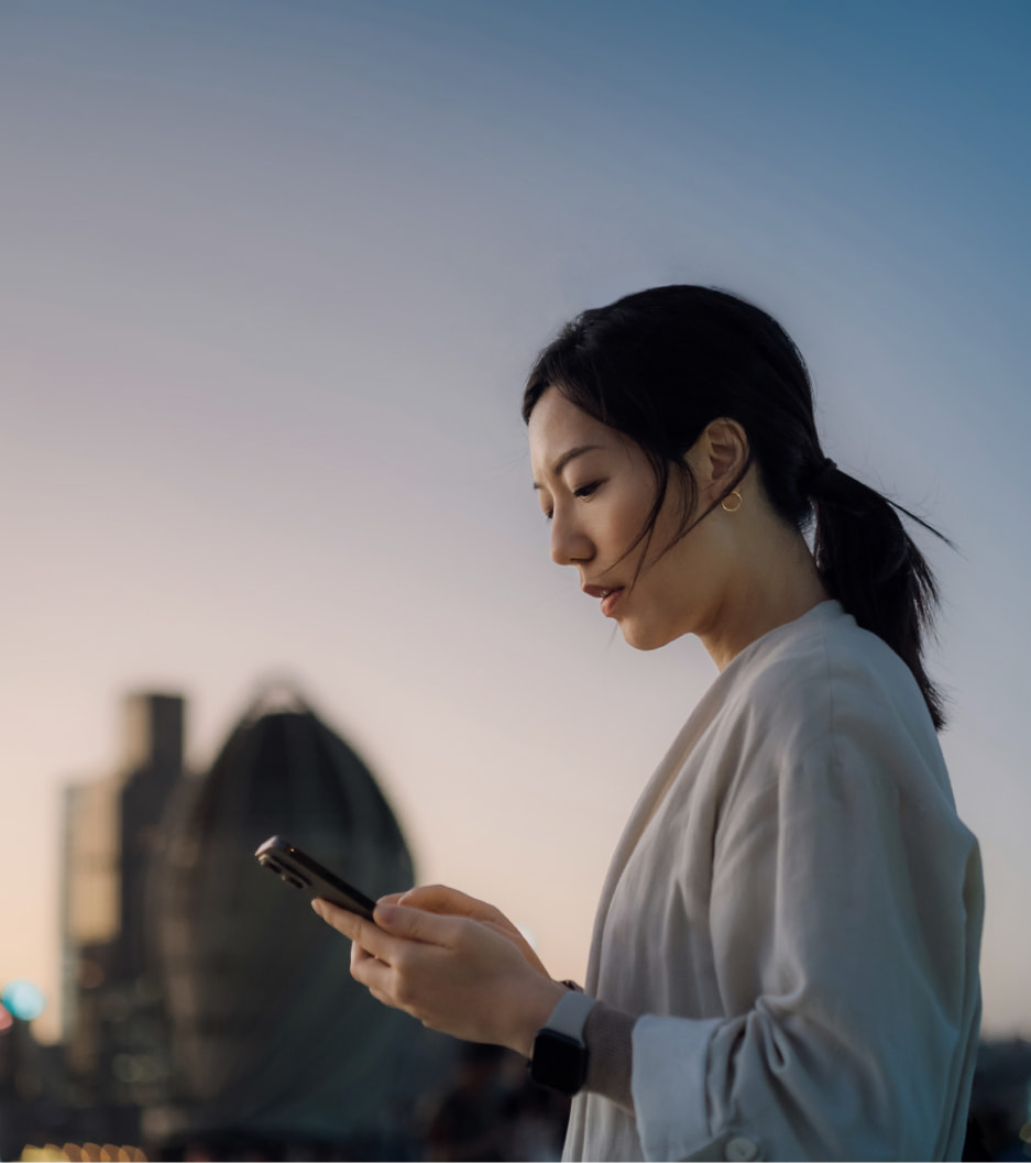Mulher com casaco claro observa o celular, com prédios ao fundo e luz do entardecer.