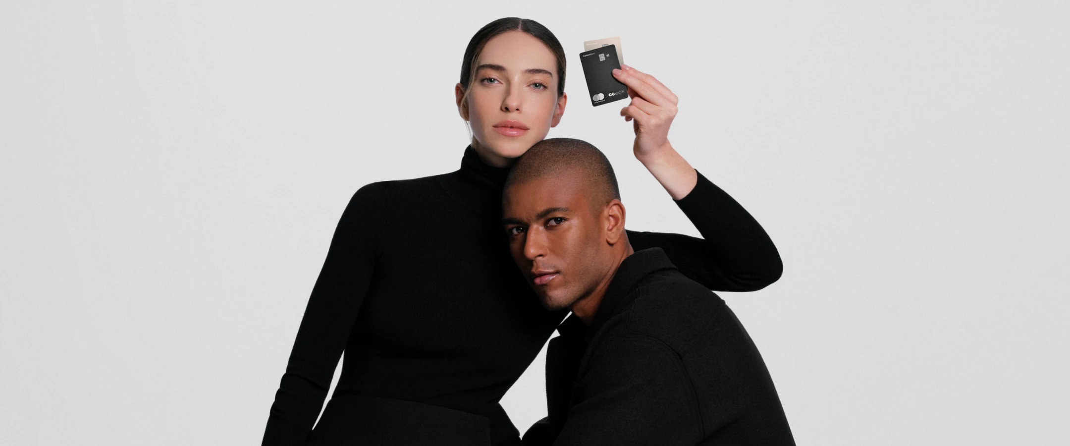 Mulher segura um Cartão C6 Carbon Mastercard Black enquanto um homem se apoia nela. Ambos vestem preto.
