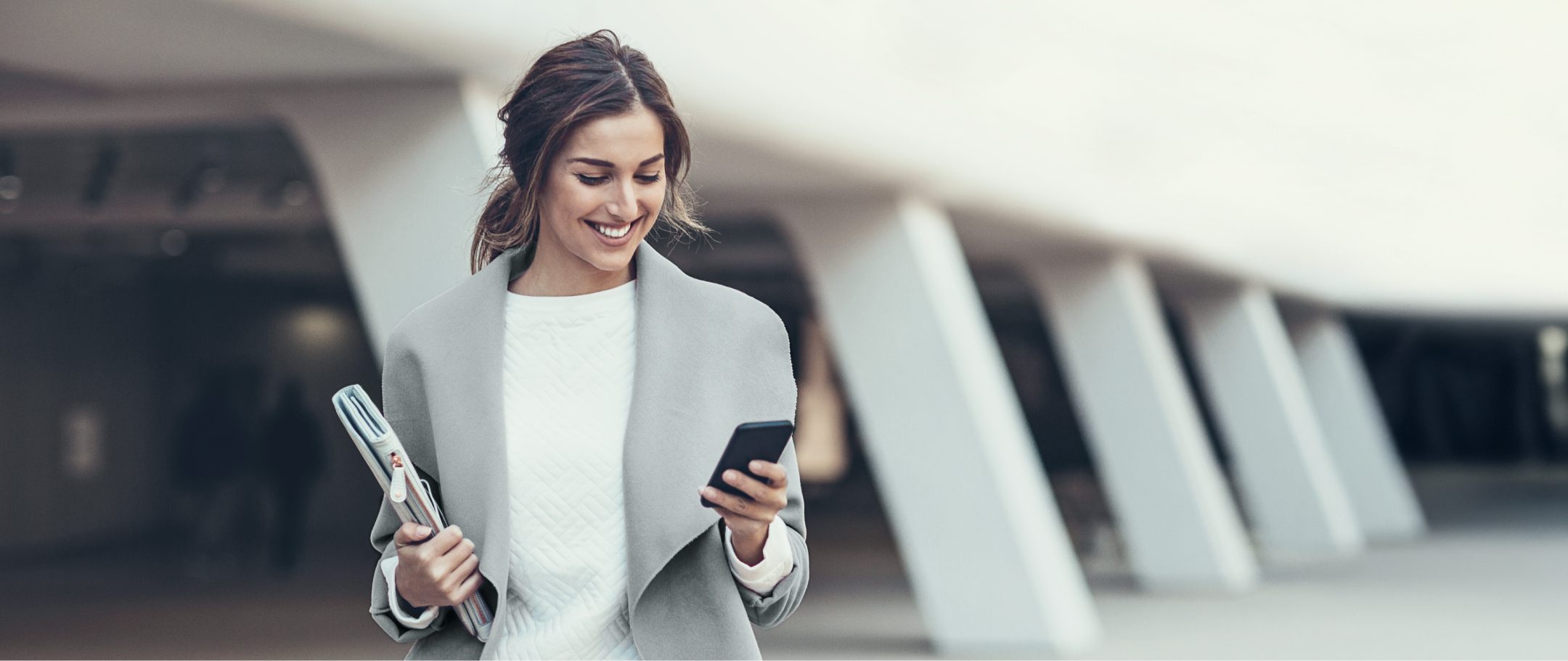 Mulher sorrindo enquanto caminha com celular e pasta em ambiente urbano.