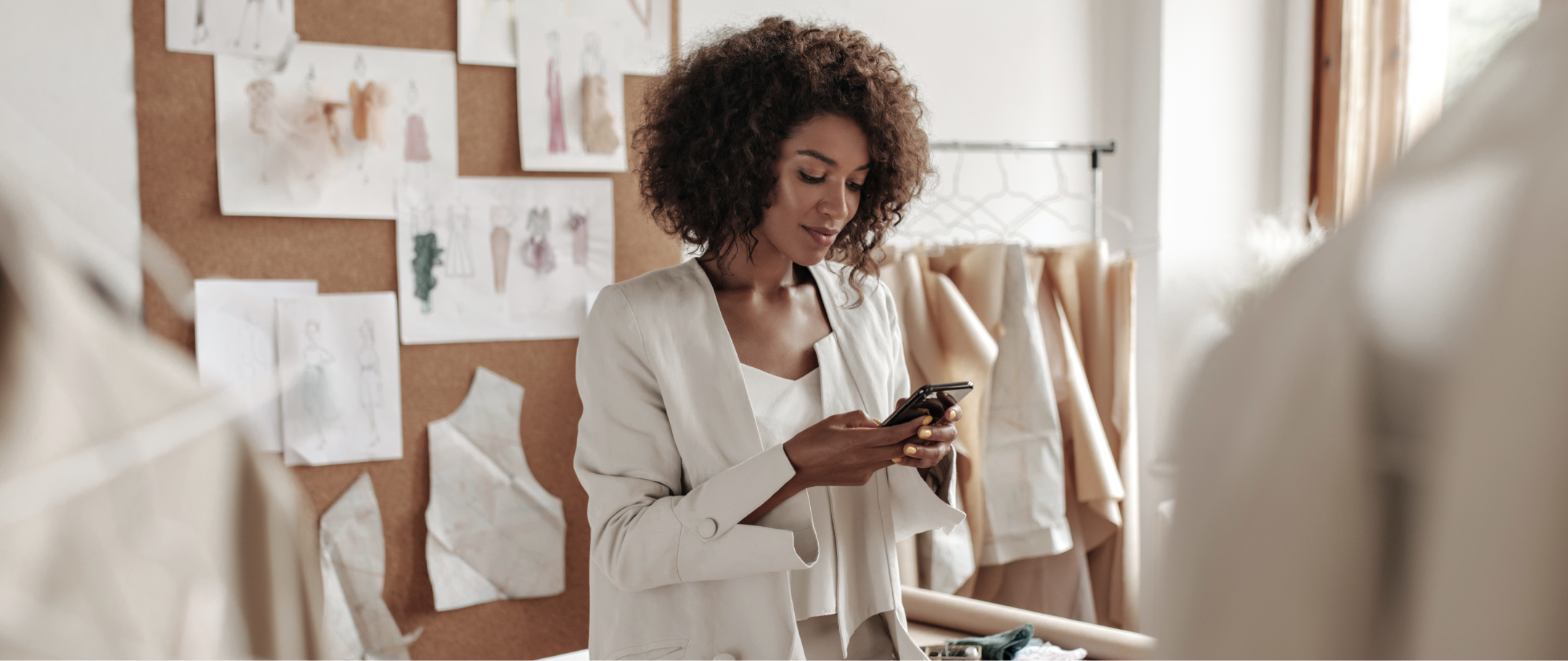 Mulher negra, de terno branco, mexendo no celular em meio a um ateliê