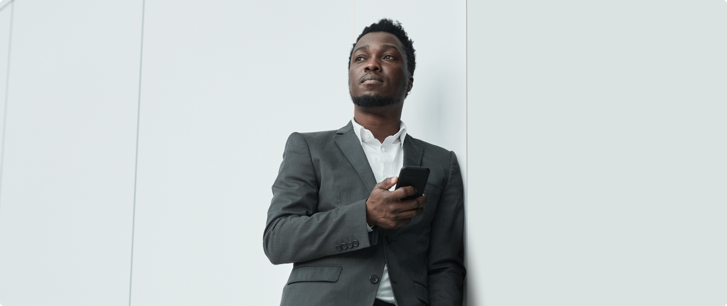 Foto de homem negro encostado na parede. Homem veste blazer e camisa branca e segura um celular na mão direita.