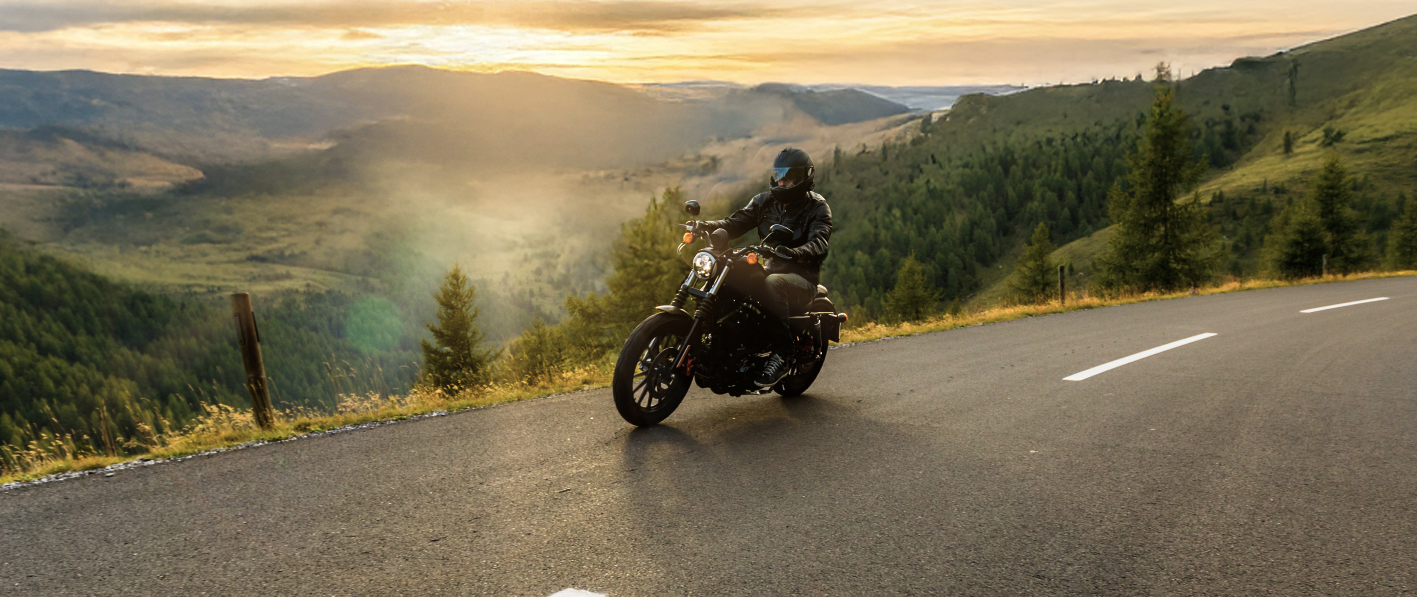 Motociclista em estrada de serra ao entardecer, com montanhas verdes ao fundo e céu alaranjado.
