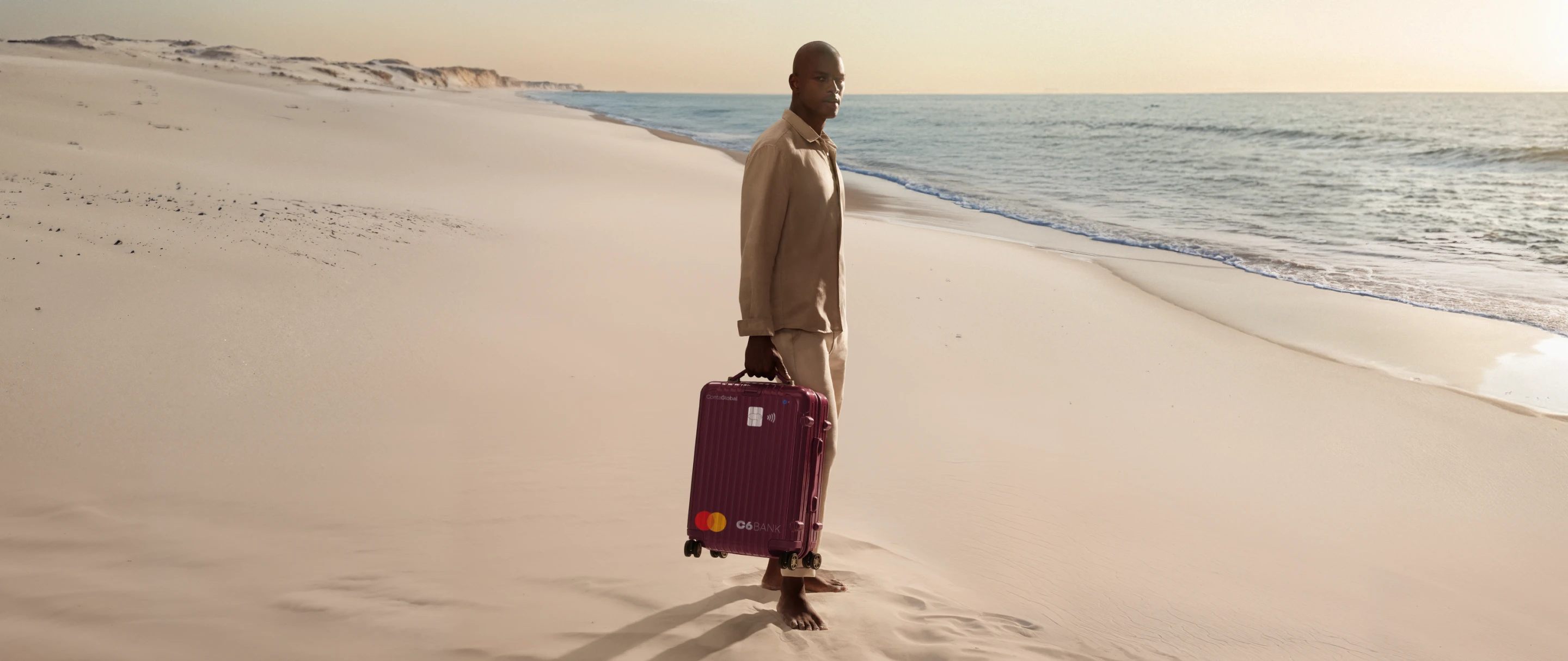 Homem na praia puxa mala com layout do C6 Conta Global. Cena ao entardecer transmite mobilidade e tranquilidade.