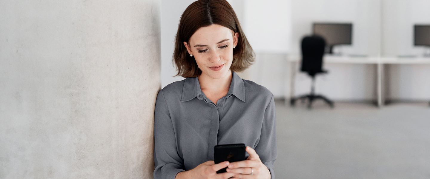 Mulher encostada na parede, olhando para o celular e sorrindo em um ambiente de escritório.