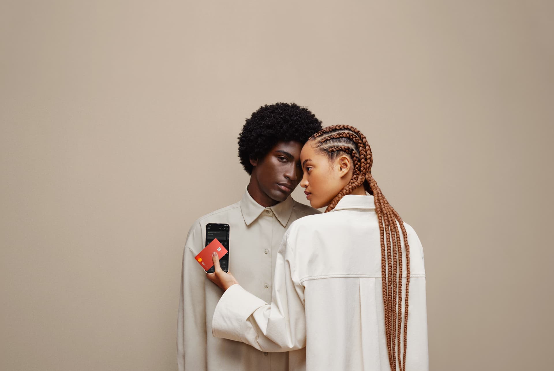 A imagem mostra um homem e uma mulher negros. A mulher está de frente para o homem, mas de costas para a camêra e olhando para a esquerda. Nas mão estão um celular e o cartão C6 Red 