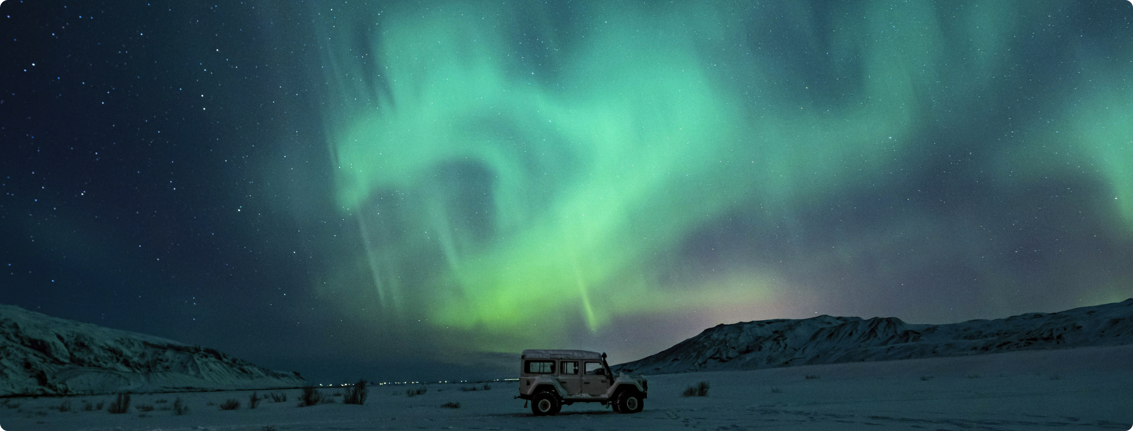 Aurora boreal verde no céu estrelado sobre neve, com veículo parado e montanhas ao fundo.