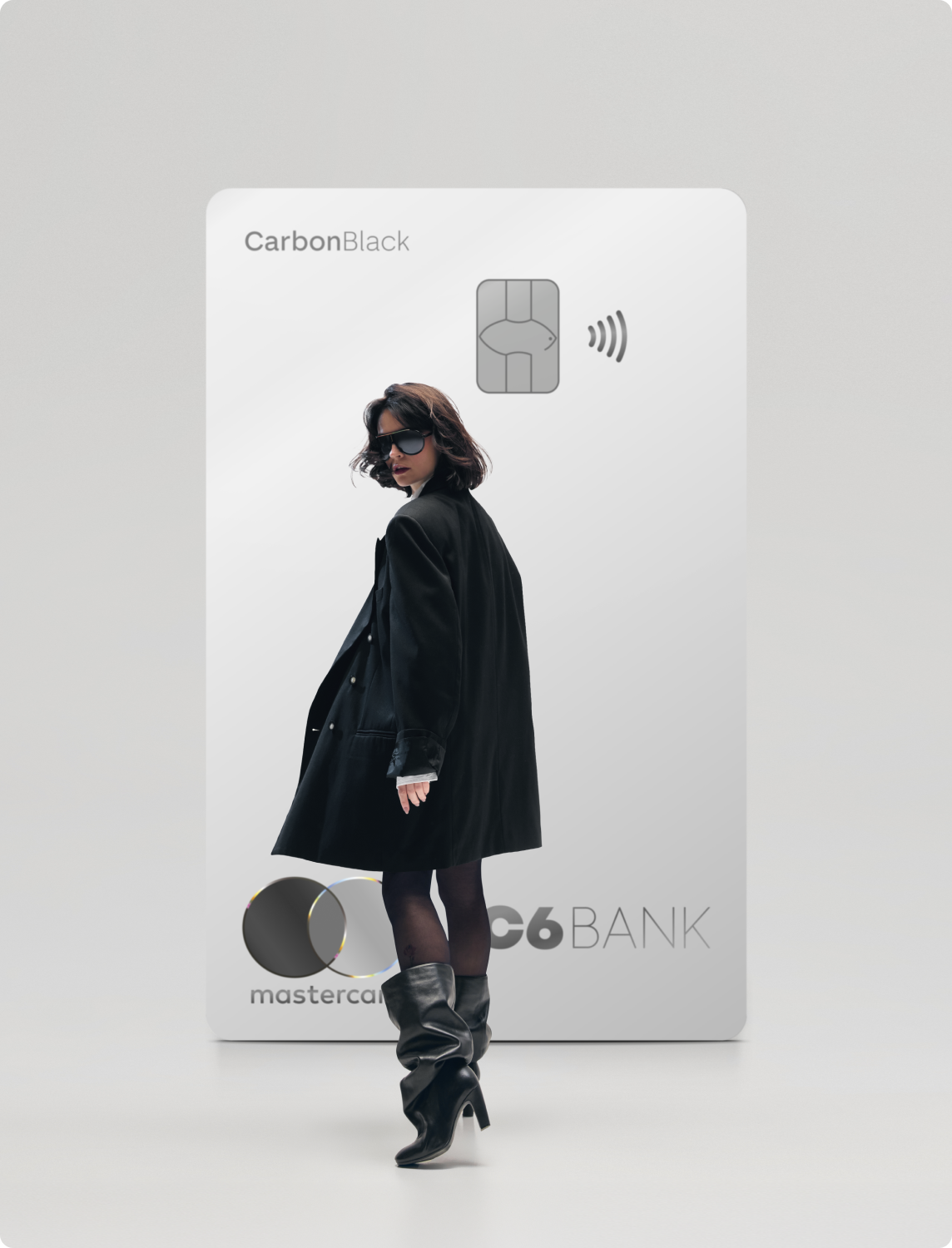 Mulher de preto caminha à frente de um Cartão C6 Carbon Mastercard Black prata em tamanho ampliado, sobre fundo claro.