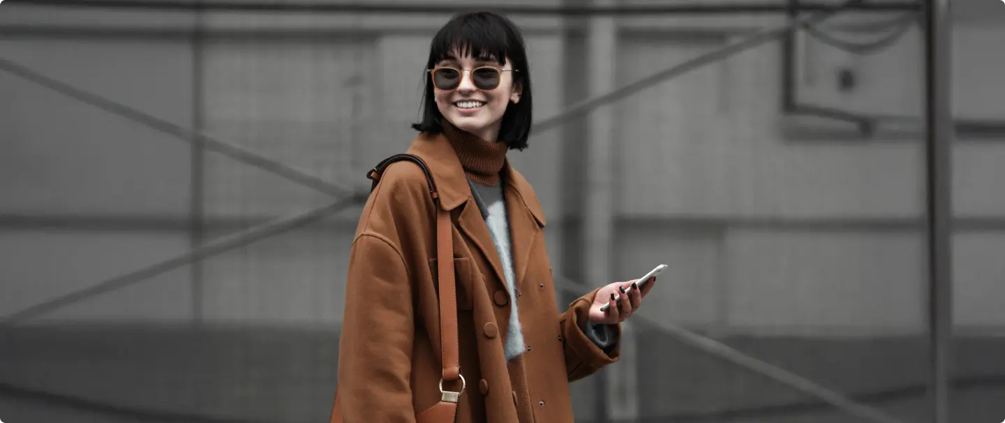 Mulher sorrindo com celular na mão Foto de mulher sorrindo com cabelo curto preto e óculos de sol marrom segurando um celular com a mão esquerda.