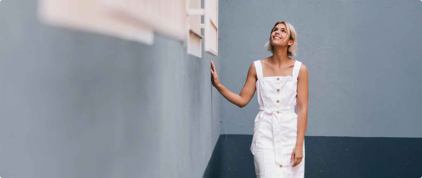 Mulher ao lado de casa azul Fotografia de mulher com vestido branco e cabelo loiro, apoiada em uma parede azulada.