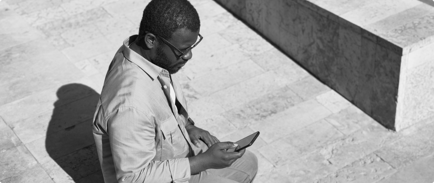 Foto em preto e branco de homem negro sentado segurando um celular com a mão direita