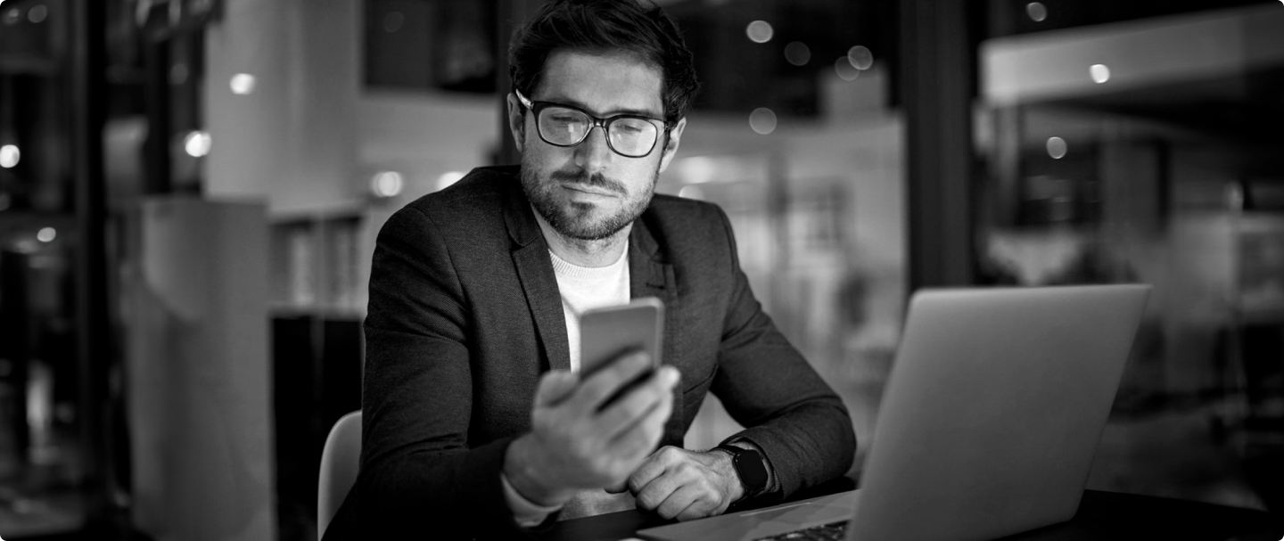 Imagem em preto e branco de homem de paletó e óculos sentado olhando para o celular em sua mão