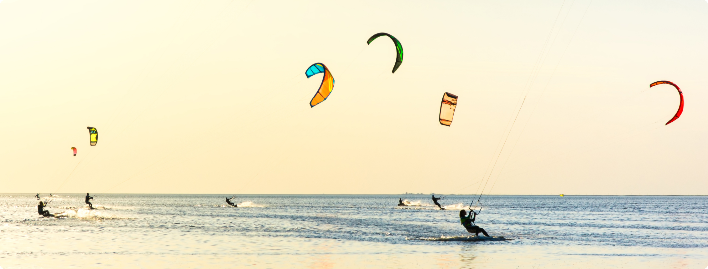 Pessoas fazem kitesurf com pipas coloridas sobre o mar ao entardecer.