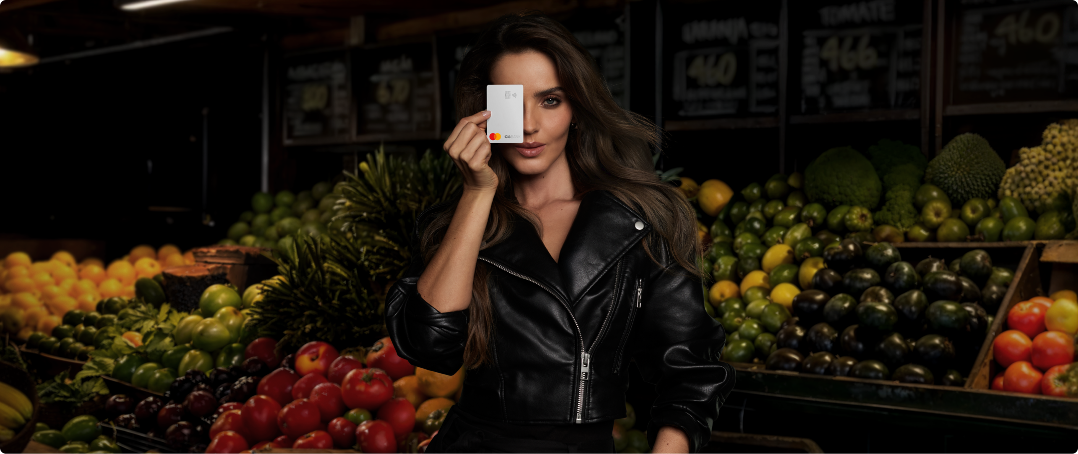 Mulher com cartão em feira SHOOTING 25-26 Mulher cobre um olho com Cartão C6 na cor Silver em feira iluminada, cercada por frutas. Cena com forte contraste.