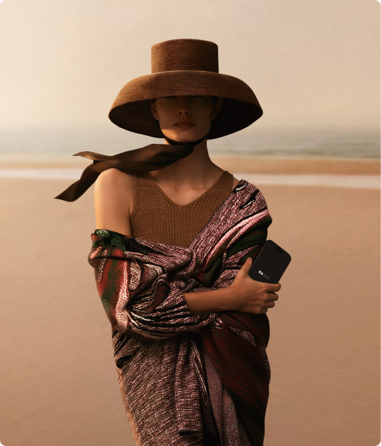 Mulher em pé na praia com mar ao fundo, usando chapéu de abas largas e roupa colorida, segurando celular junto ao corpo.