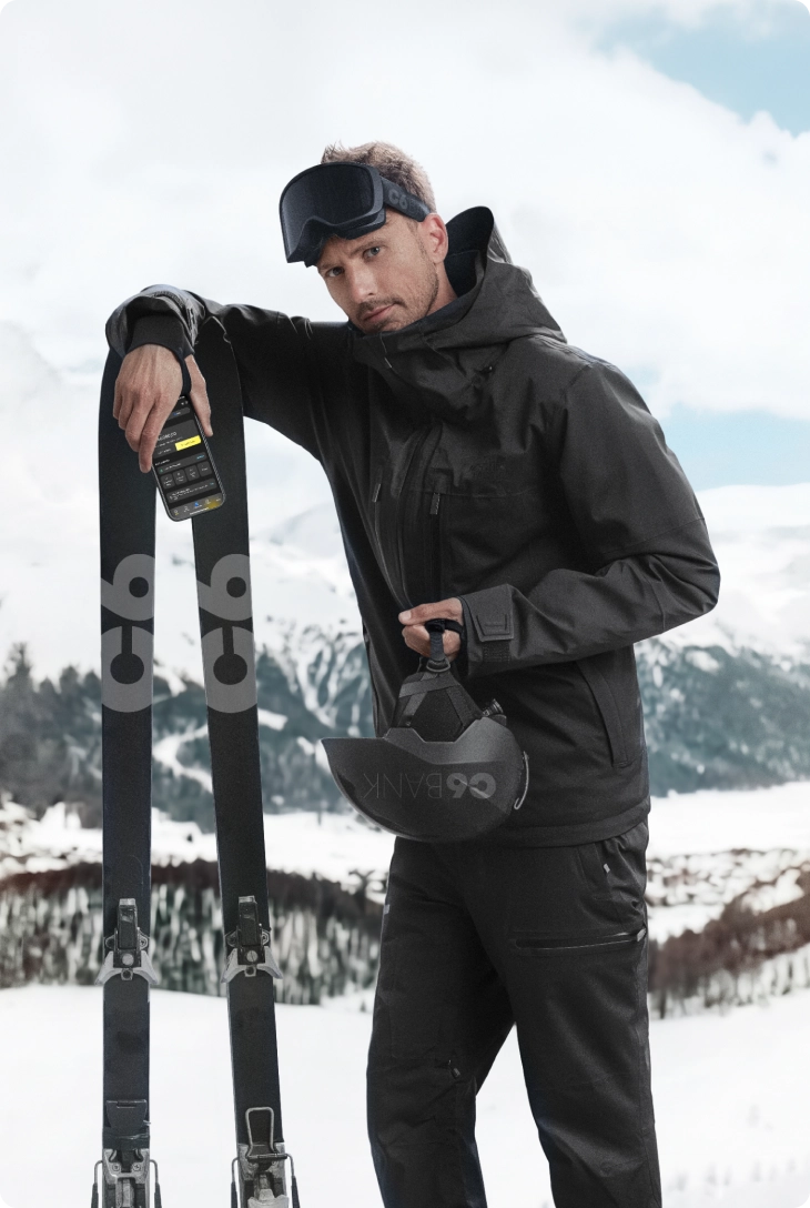 Homem em paisagem de neve com montanhas ao fundo, usando roupa de inverno, apoiada em esquis, segurando capacete, com celular exibindo app do C6 Bank, posicionado sobre o equipamento.