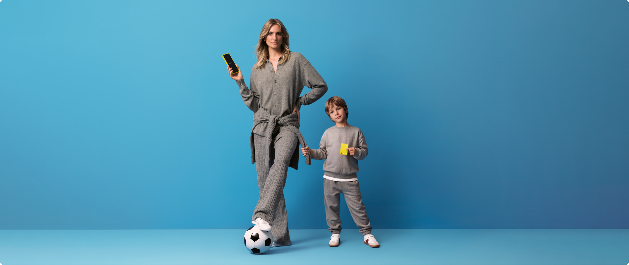 Mulher com criança em fundo azul C6 Yellow S 26-27 Mãe e filho em fundo azul. Mulher adulta segurando celular exibindo o app C6 Bank e com bola de futebol apoiada em um dos pés. Ao lado, criança segurando cartão C6 Yellow na cor amarela,