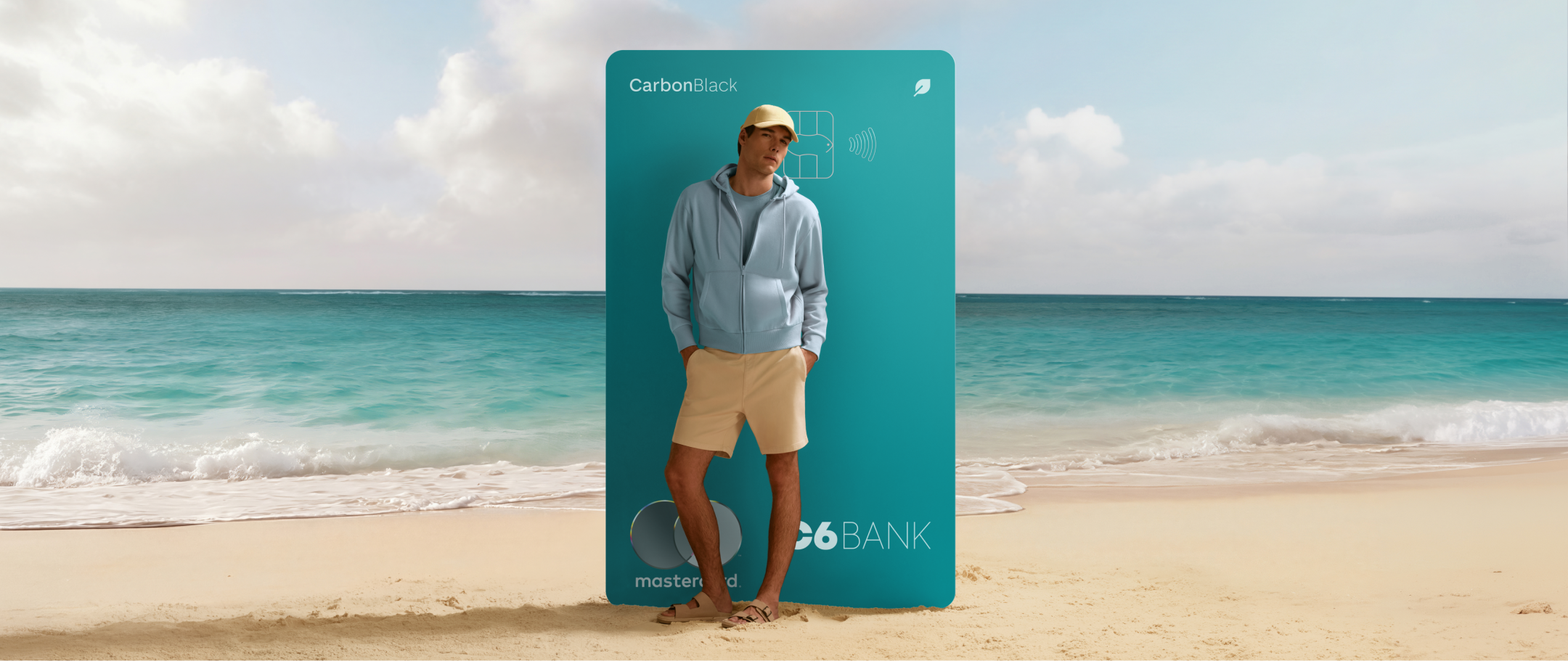 Homem apoiado em cartão na praia S 26-27 Homem em pé na areia da praia com mar ao fundo, posicionada à frente de cartão C6 Carbon Acqua gigante, usando roupa clara.