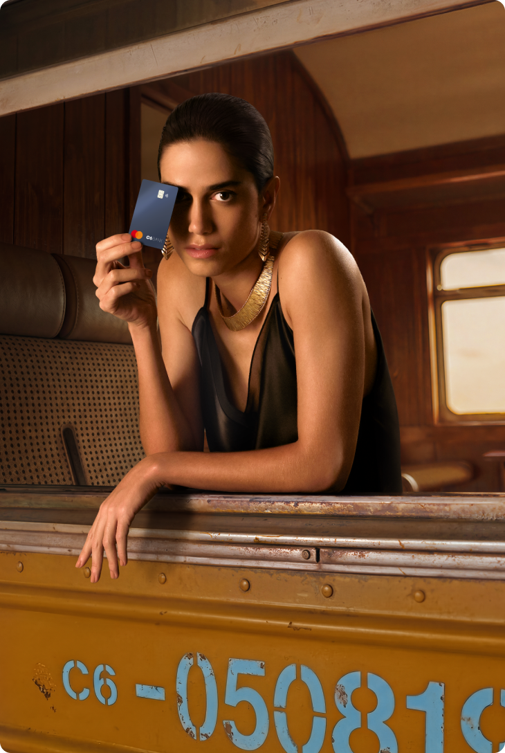 Mulher apoiada na janela de trem, com corpo inclinado para fora, segurando cartão C6 Carbon na cor preta próximo ao rosto, com interior do vagão ao fundo.