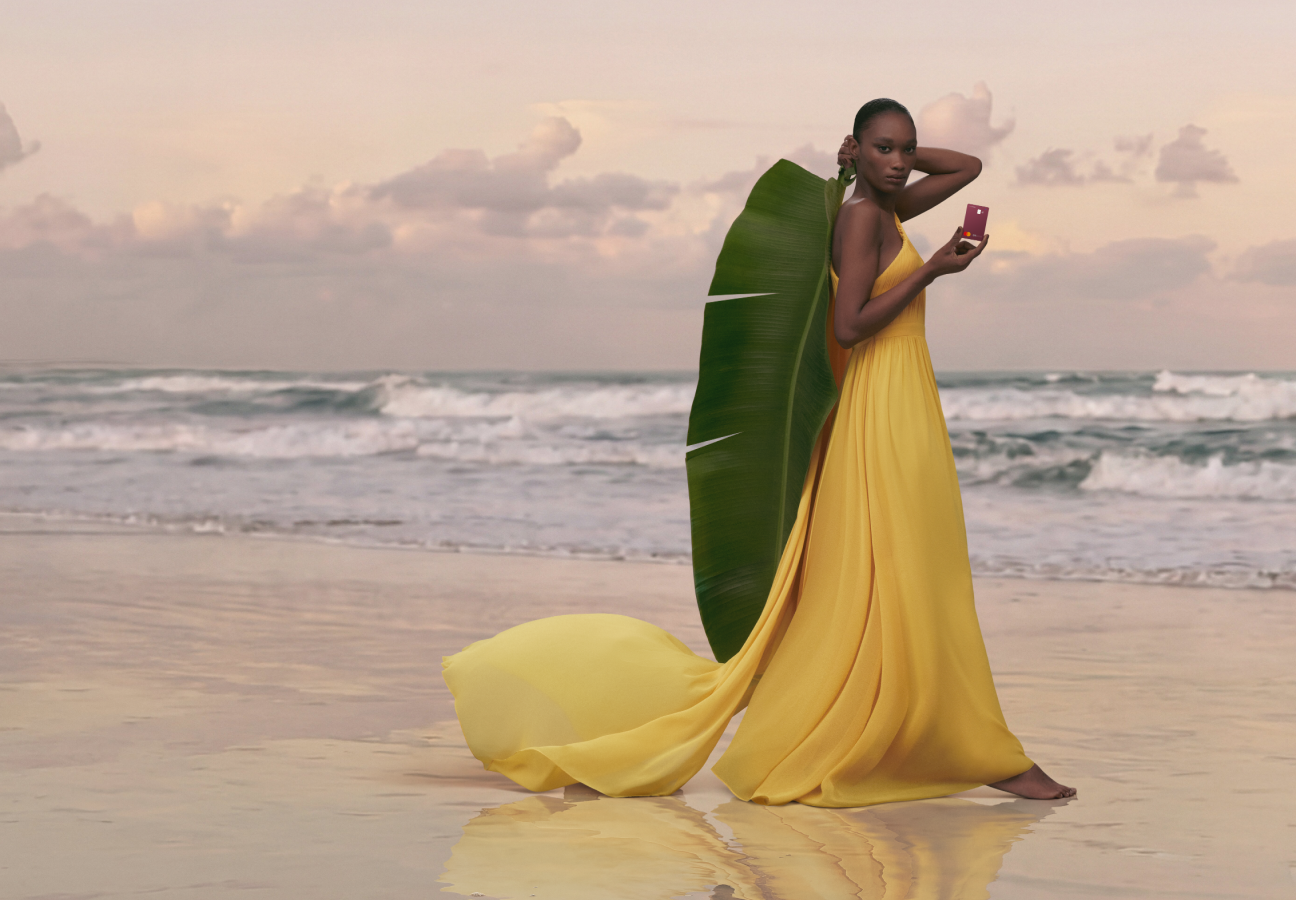 Mulher em pé na praia com mar ao fundo, usando vestido amarelo e segurando cartão C6 Conta Global na cor vinho, com grande folha verde apoiada ao lado do corpo.