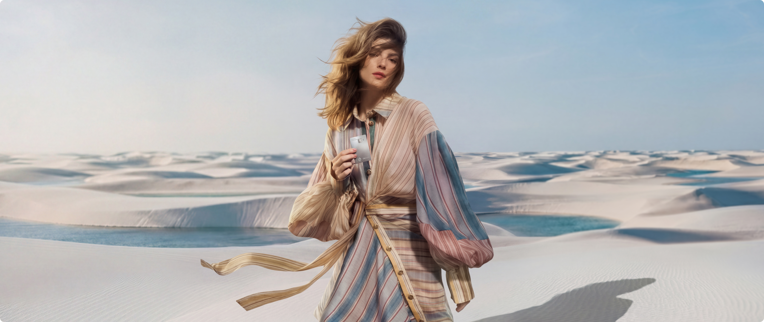 Mulher em dunas de areia com cartão S 26-27 Mulher em paisagem de dunas de areia, usando roupa listrada e segurando cartão C6 Graphene junto ao corpo, com tecido sendo movimentado pelo vento.
