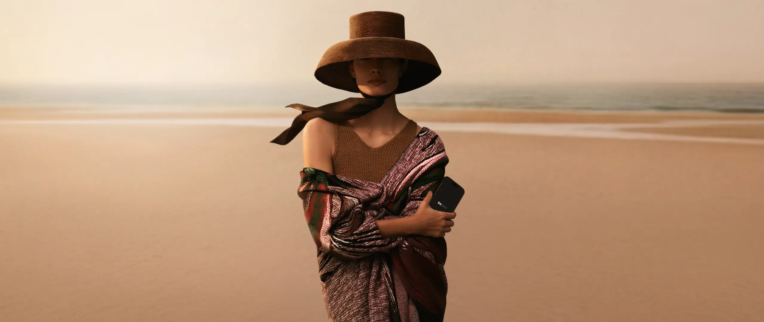 Mulher com chapéu e celular na praia S 26-27 Mulher em pé na praia com mar ao fundo, usando chapéu de abas largas e roupa colorida, segurando celular junto ao corpo.
