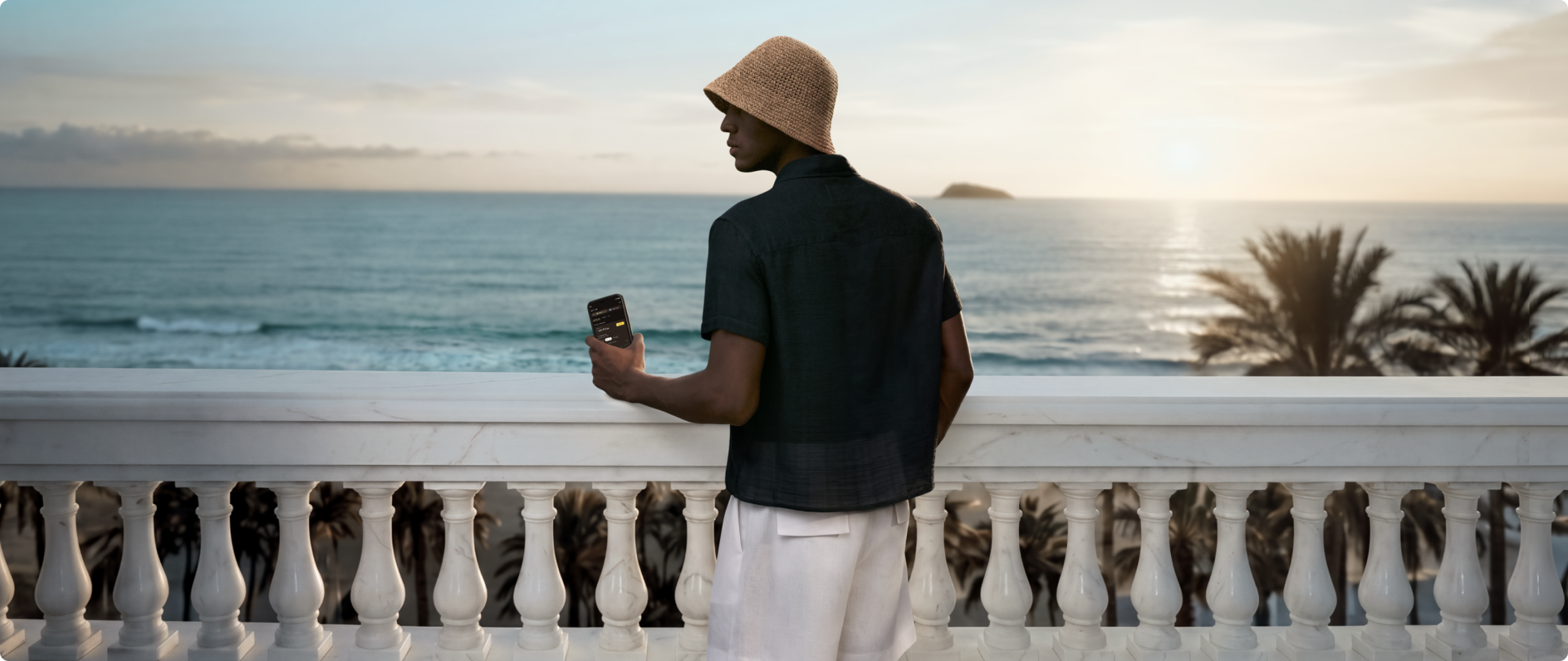 Homem em sacada de hotel beira mar S 26-27 Homem em pé em varanda com vista para o mar, usando chapéu e roupa clara, segurando celular enquanto observa o horizonte.