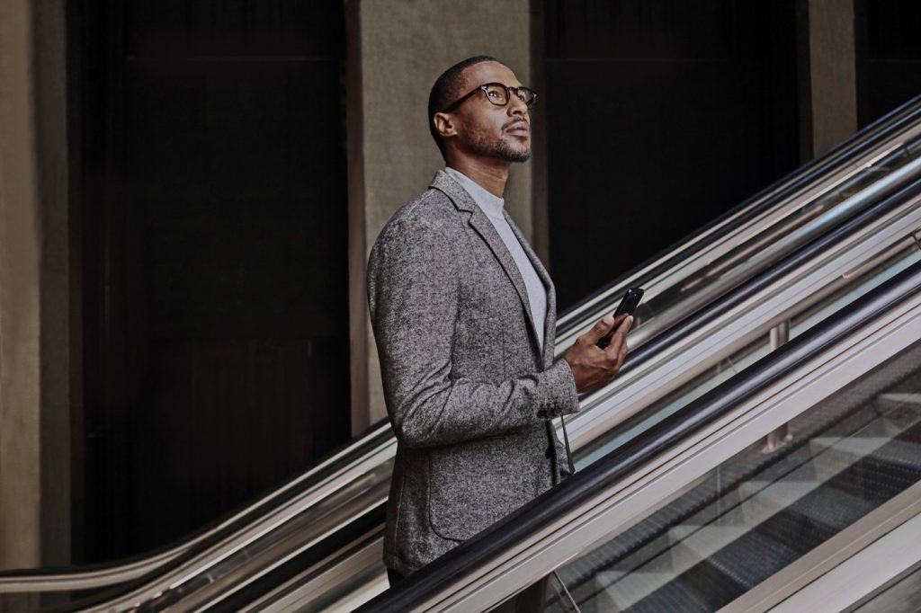 Imagem mostra homem negro de óculos e roupa cinza subindo uma escada rolante com um smartphone na mão