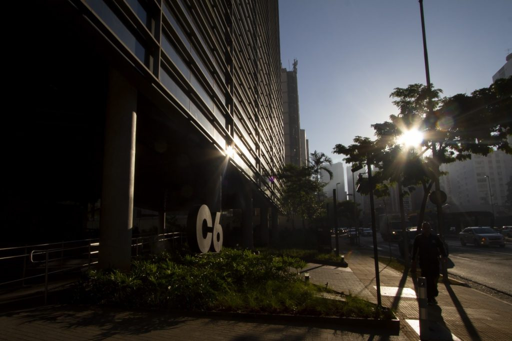 Imagem mostra a fachada do C6 Bank com tons escuros e o sol nascendo ao fundo