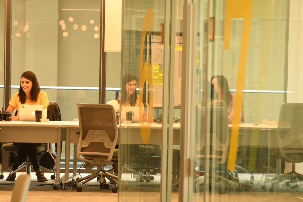 A imagem mostra um grupo de pessoas em reunião em uma sala com paredes de vidro e post-its colados