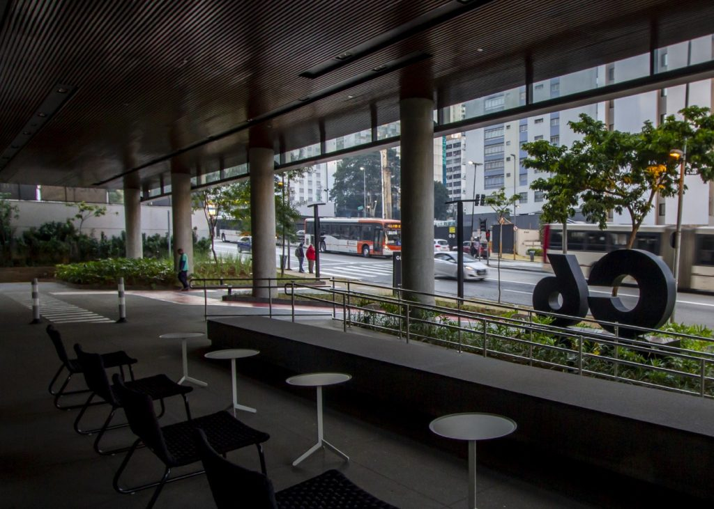 Imagem mostra a fechada do C6 Bank vista de dentro. Do lado da rua, é possível ver o movimento da avenida com pessoas e veículos e o logo do C6 Bank à direira