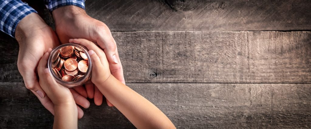 A imagem mostra as mãos de uma criança entregando um pote de moedas para um adulto