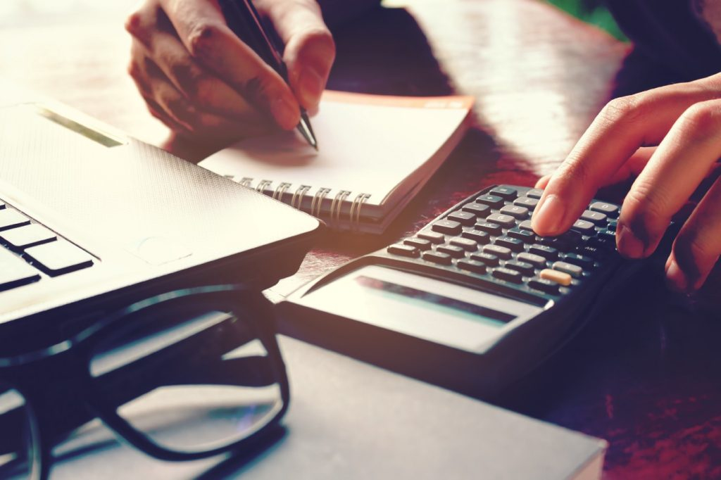 Imagem mostra uma pessoa fazendo contas com o auxílio de uma calculadora e anotando em um caderno