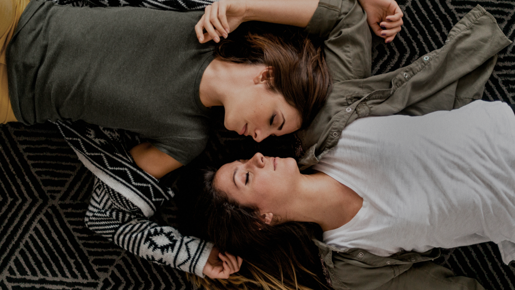 Imagem mostra duas mulheres brancas usando roupas neutras deitadas no chão e de olhando de forma apaixonada