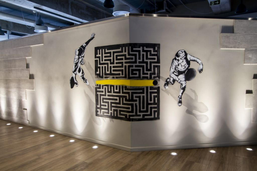 Foto de uma grafite em uma das paredes internas do prédio do C6 Bank. Nele, duas pessoas jogam frescobol e a bolinha passa por cima de um labirinto deixando um rastro amarelo.