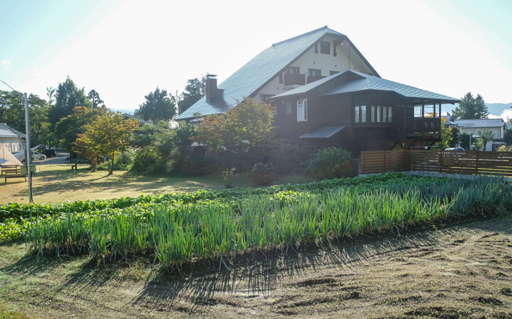 Uma casa grande em um ambiente rural, com um jardim próximo, incluindo plantações de cebolas no primeiro plano e árvores ao fundo, sob um céu claro.