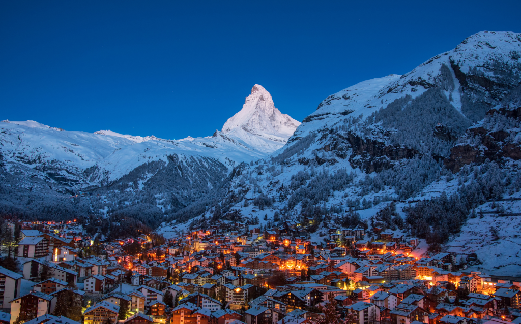 Vista noturna da cidade de Zermatt com o Matterhorn ao fundo, coberto de neve e iluminado. Um cenário mágico nos Alpes Suíços.
