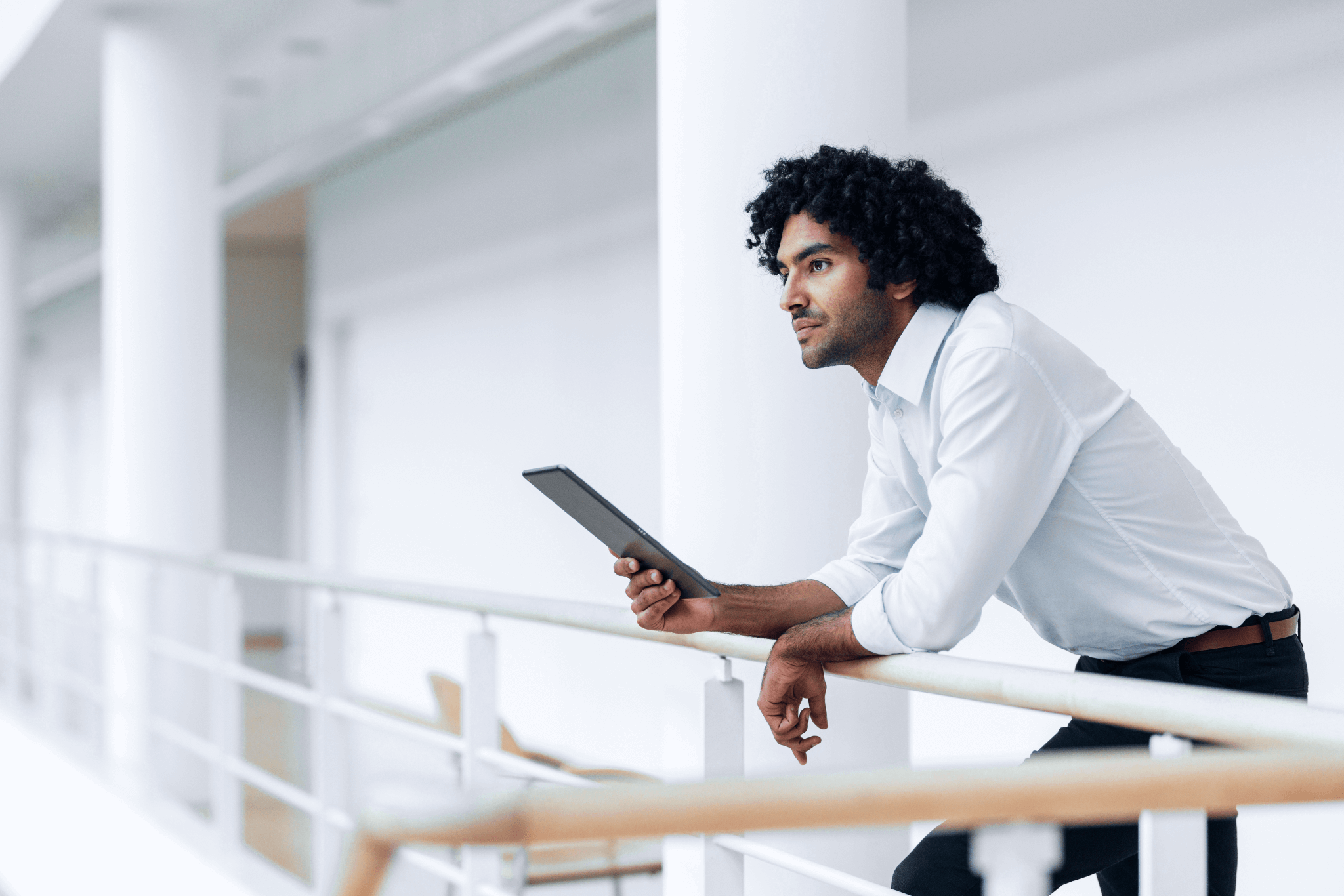 Um homem com cabelo cacheado, vestido de camisa branca, olhando pensativo enquanto segura um tablet na mão, em um ambiente moderno e minimalista.